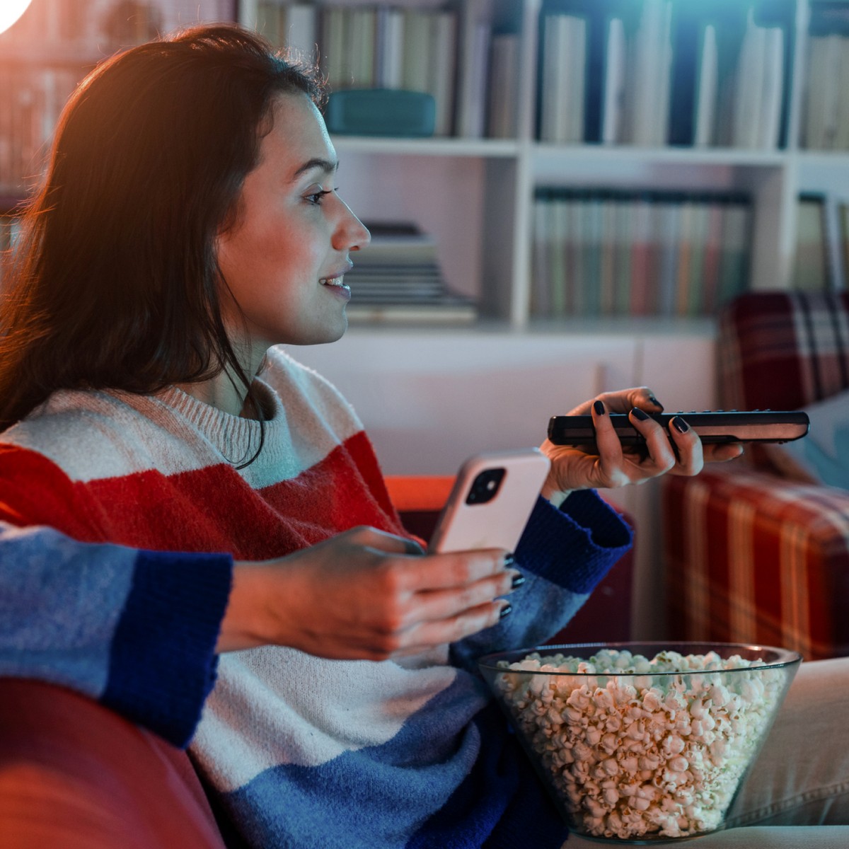 A woman sitting on the couch holding her phone and a tv remote with a bowl of popcorn